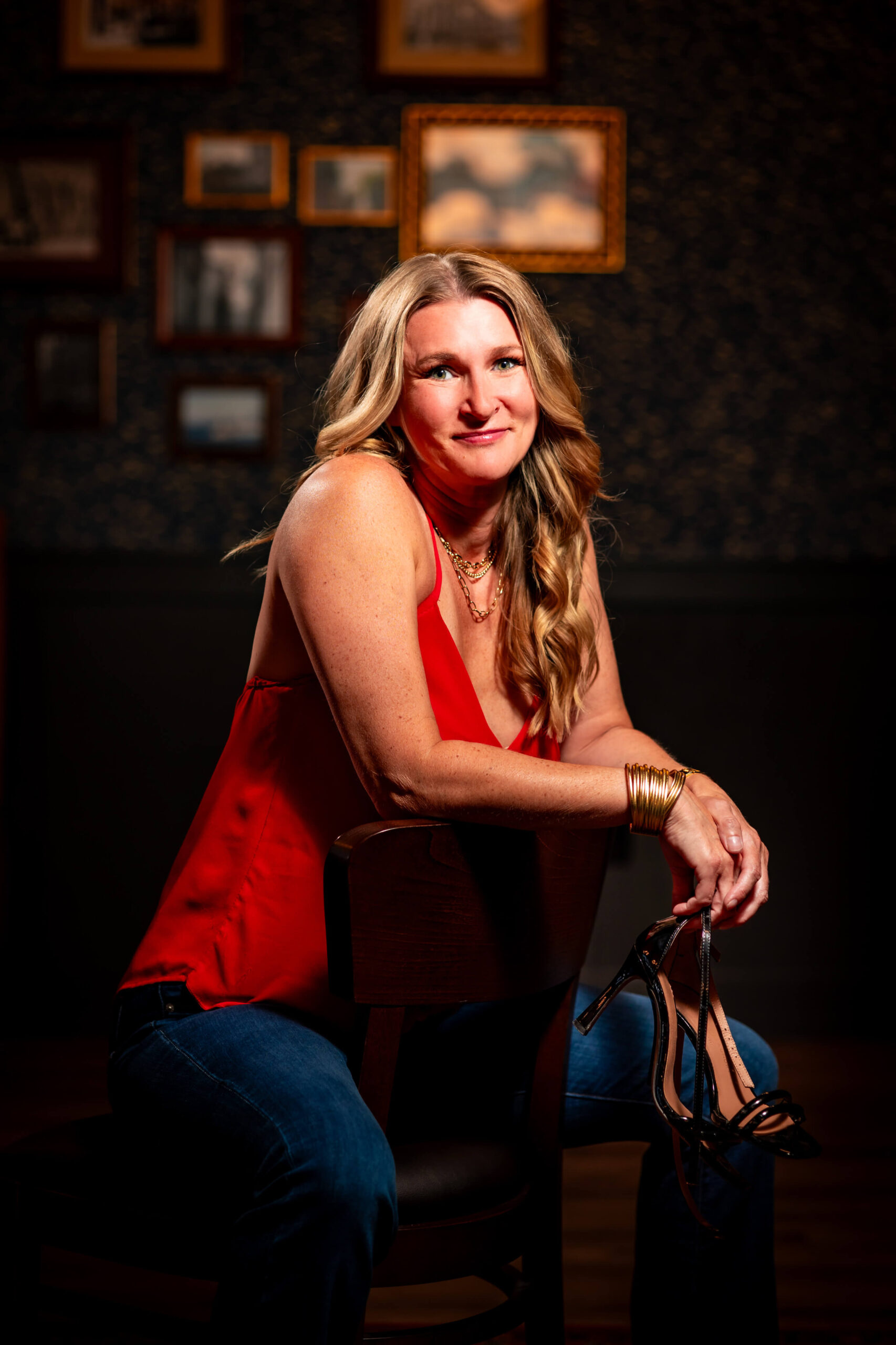 Callie Croken, a life and business coach, sitting in a relaxed and reflective pose in front of unique wall frames during her branding photoshoot with Lake Design holding her high heels in her hand.